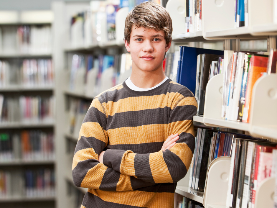 teenage boy in yellow and gray striped shirt leaving against library shelves full of books